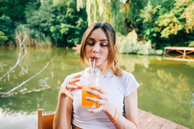 Genç ve çekici bir kadın portakallı limonata içiyor, açık denizde bir iskelede dinleniyor.