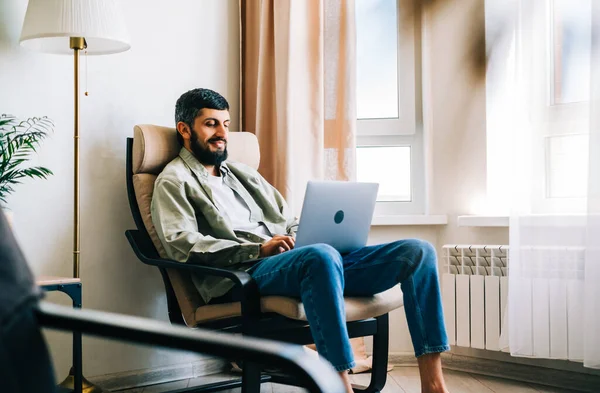 Caucasian man freelancer working at home, sitting on armchair and using laptop computer.