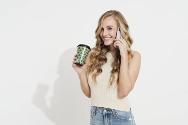 Beyaz bir duvarda gülümseyen genç sarışın kız kahve içiyor ve cep telefonuyla konuşuyor. Kameraya bakıyor.