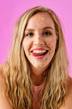 Pretty blonde girl smiling showing her tongue with colorful stickers on her face on a pink background