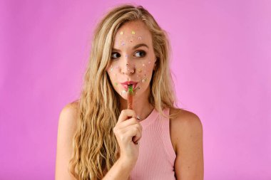 Pretty blonde girl with stickers on her face licking a candy on a pink background