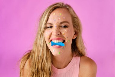 Pretty blonde girl with stickers on her face biting a candy on a pink background