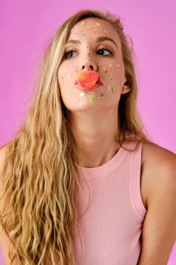 Pretty blonde girl with stickers on her face with a heart shaped candy in her lips on a pink background