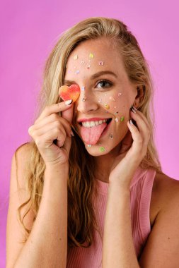 Pretty blonde girl with stickers on her face holding a heart-shaped candy in her hand covering her eye and showing the tongue on a pink background