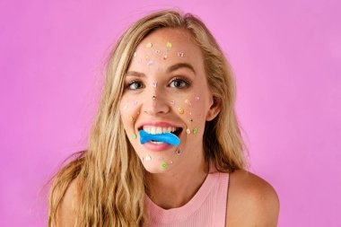 Pretty blonde girl with stickers on her face biting a candy on a pink background