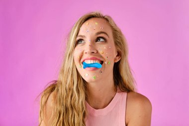 Pretty blonde girl with stickers on her face biting a candy on a pink background