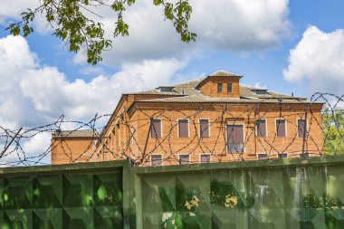 Taş hapishane binası, yeşil çit ve dikenli teller mavi gökyüzüne karşı bulutlu