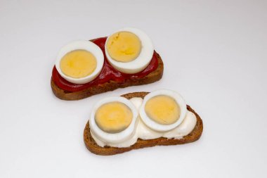 Beyaz arka planda haşlanmış yumurtalı iki çavdar ekmekli sandviç. Biri domates soslu, diğeri beyaz soslu.