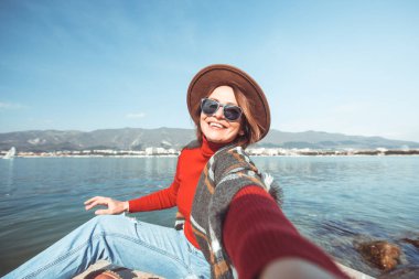 Şapkalı, genç ve güzel bir kadın deniz kenarında bir iskelede oturur ve selfie çeker..