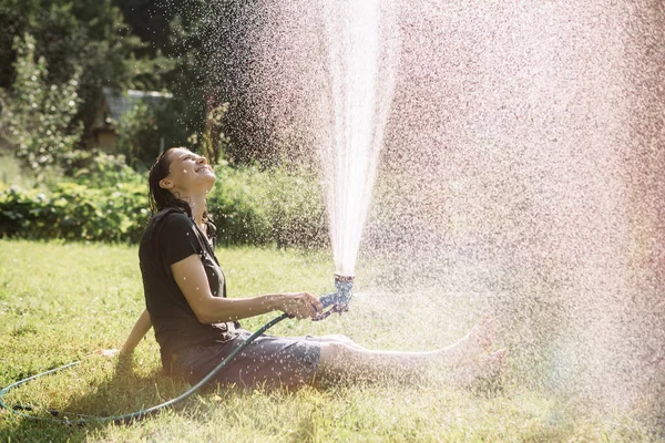 Молодая женщина сидит на траве радуется и поливает себя садовым шлангом. — стоковое фото