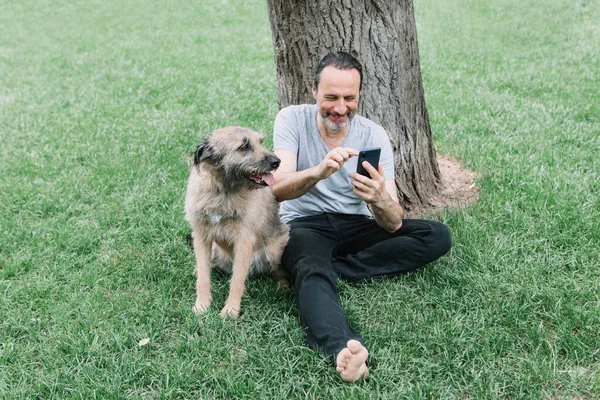 Dospělý muž středního věku se psem pod stromem na trávě komunikuje po telefonu. — Stock fotografie