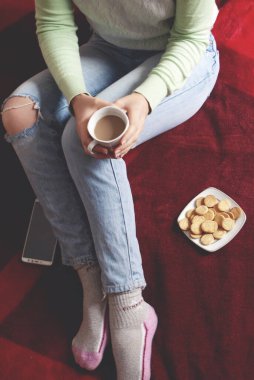 Kırmızı bir yatak örtüsünün üzerinde oturan bir kadın elinde bir fincan kahve tutuyor. Yanında beyaz bir fincan tabağında küçük kurabiyeler var..