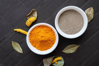 Beyaz kaselerde zerdeçal tozu ve karabiber tozu, zerdeçal kökü ve körfez yaprakları kâselerin yanında. Mevsimler üst görünüm.