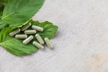 Kahverengi ahşap yüzeydeki kapsüldeki bitkisel ilaçlara seçici bir şekilde odaklan, arka planda yeşil fesleğen yaprağı bitkisi olsun. Sağlık ve tıbbi yaşam tarzı kavramı.