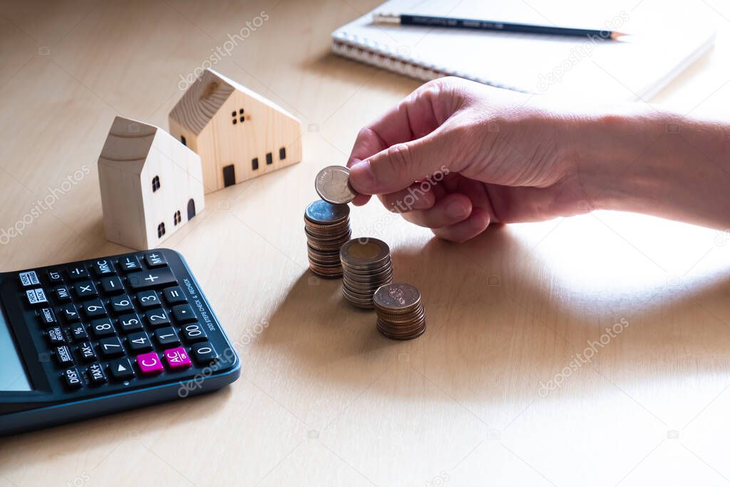 Selective focus at men hand put coin into increase stack. With blurred calculator and house mock up on wooden desk. Personal financial saving concept for home loan, mortgage investment.