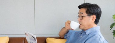 Bayrak atışı. Yüze seçici odaklanma. Asyalı kıdemli adam sabahları kahve içerken kanepede makara yapıyor. Rahatlama yaşam tarzı ve rutin ile mutlu emeklilik.