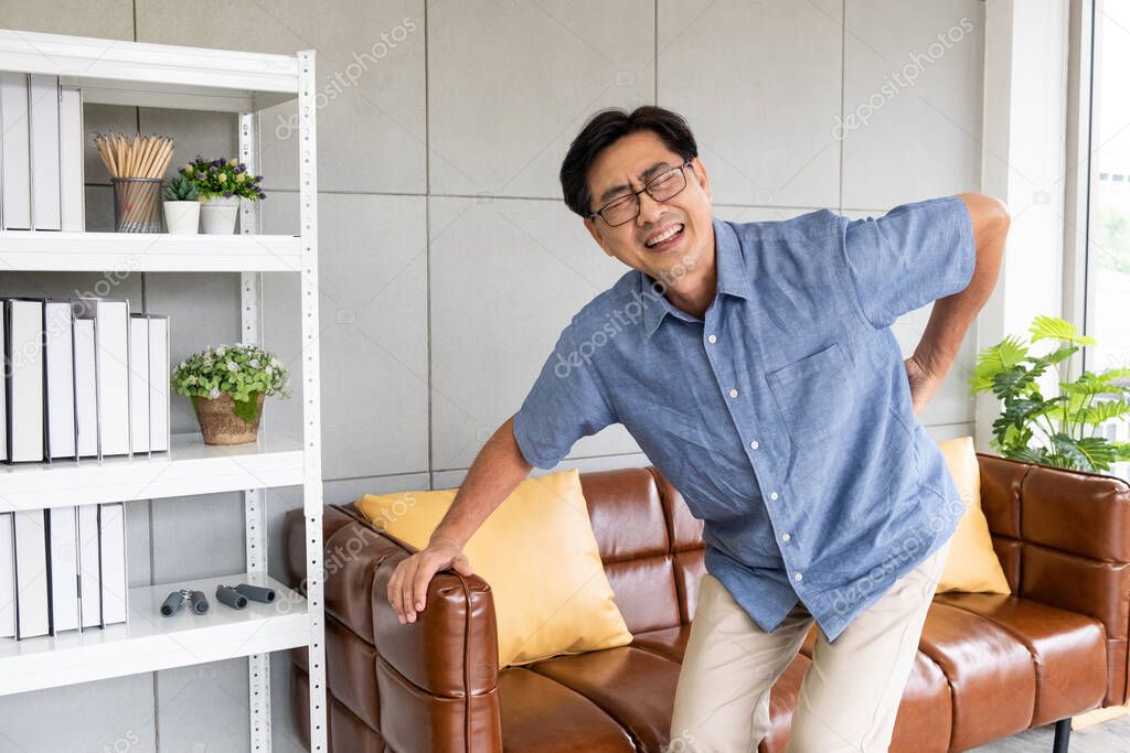 Indoor shot of senior Asian men sit on the sofa couch and touch his back and suffering from back pain, aching muscles, bones or lumbago. Elderly health problem concept.