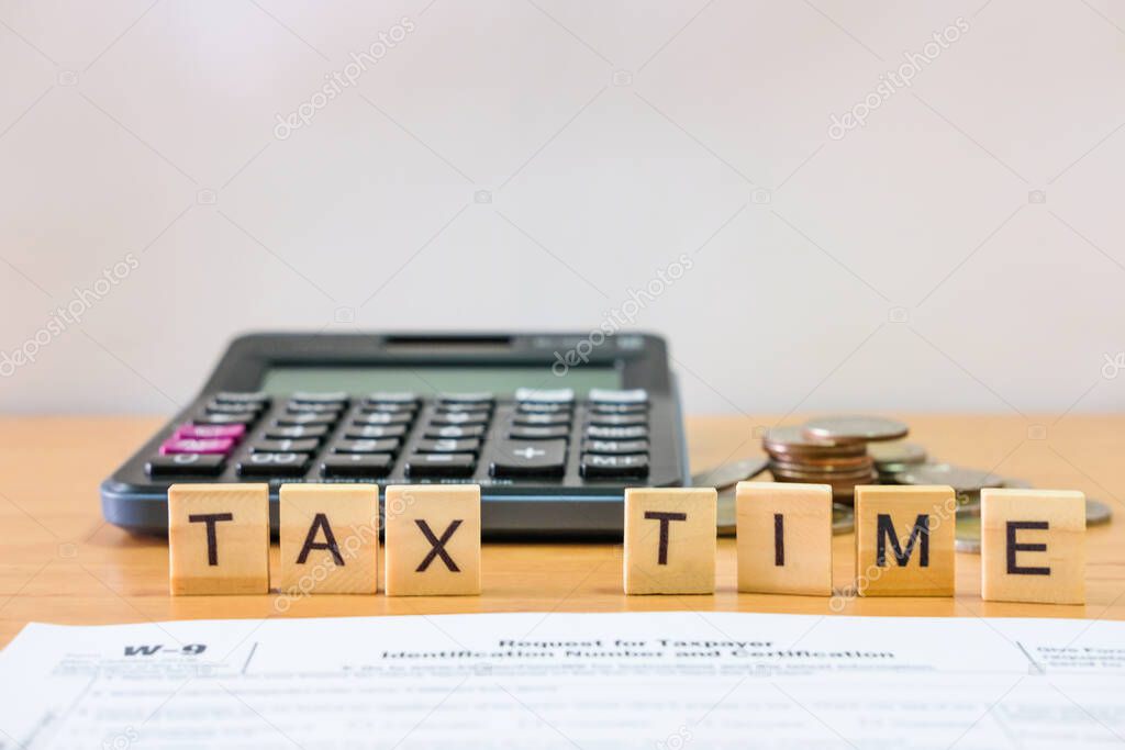 Selective focus at wood tax time sign with blurred calculator and coins stack and white copy space background. Business or personal tax payment, expense that need to manage and plan.