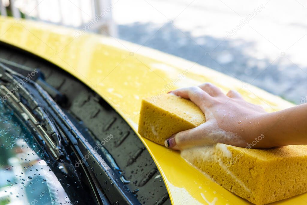 Selective focus at women hand using sponge to wash and clean colorful yellow car with soap bubble at home. Self car care maintenance activity.