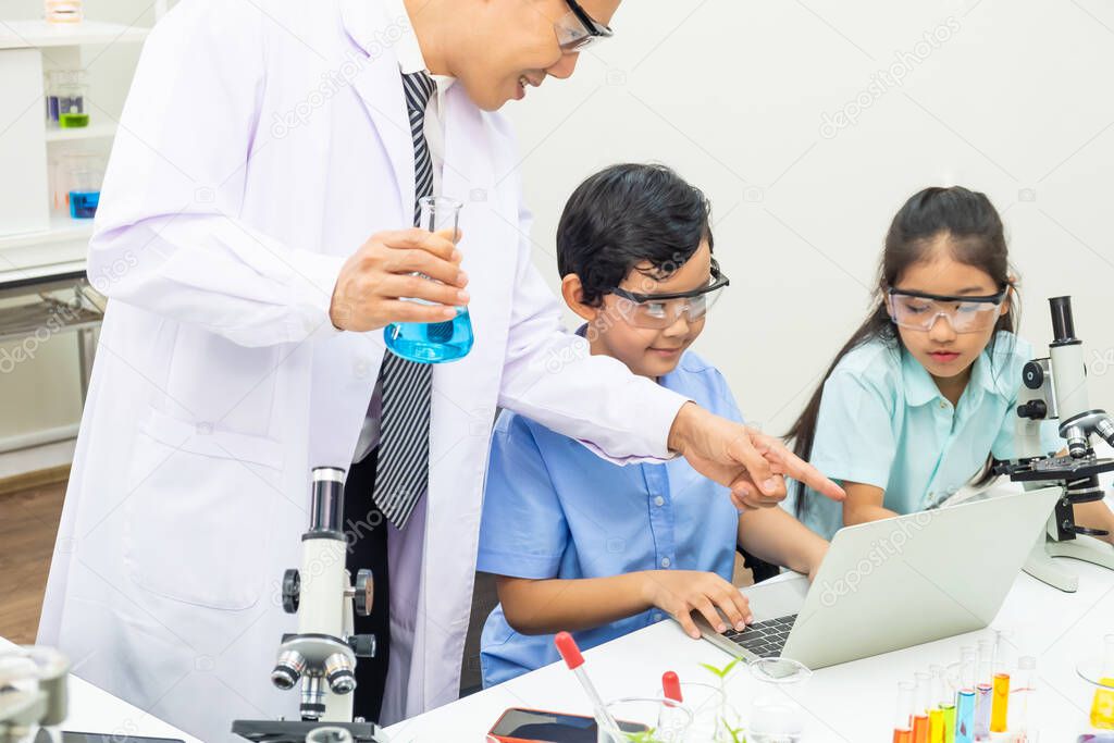 Young Asian boy and girl student wearing protective google while study and do science experiment in classroom with teacher. Learning and having fun moment. Chemist or biology classroom concept.