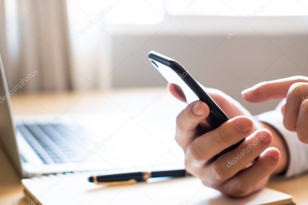 Selective focus at Smartphone. Men hand holding mobile phone while using finger to touch device screen. With blurred computer laptop and paper notebook with pen on working desk. Wireless technology.