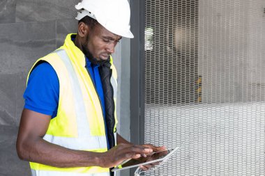 İnşaat alanındaki Siyah Afrikalı ustabaşının yüzüne seçici odaklanma, bilgi kaydetmek için dijital tablet kullanırken koruyucu şapka ve güvenlik ekipmanı takıyor. İnşaat mühendisi çalışıyor.