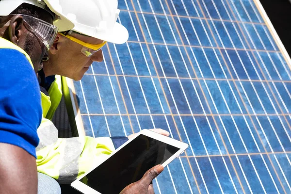 Selective focus at digital tablet. African and Caucasian engineer inspect electrical solar panel and record information into mobile tablet. Alternative energy and industrial concept. Outdoor shot.