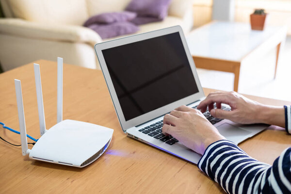 Selective focus at men hand while type on computer laptop keyboard while using internet. In front of high speed wireless internet router that provide Wi-fi signal inside of the house. 