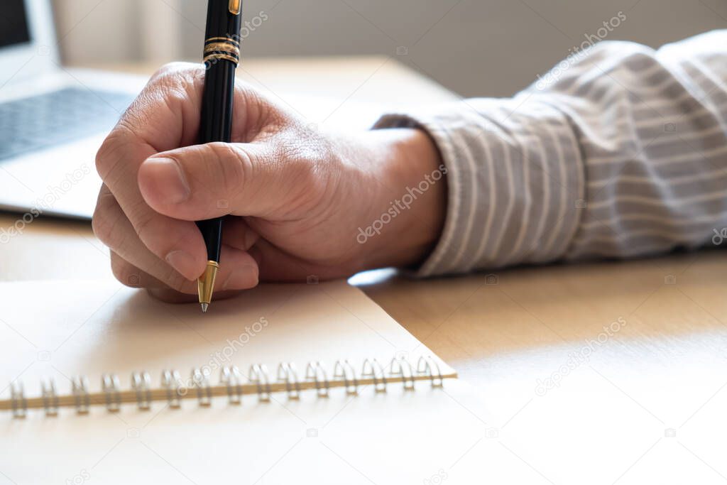 Selective focus at human hand using pen to write down short note on paper on working table. With blurred laptop computer at the background. List for plan or organize activity during working days.