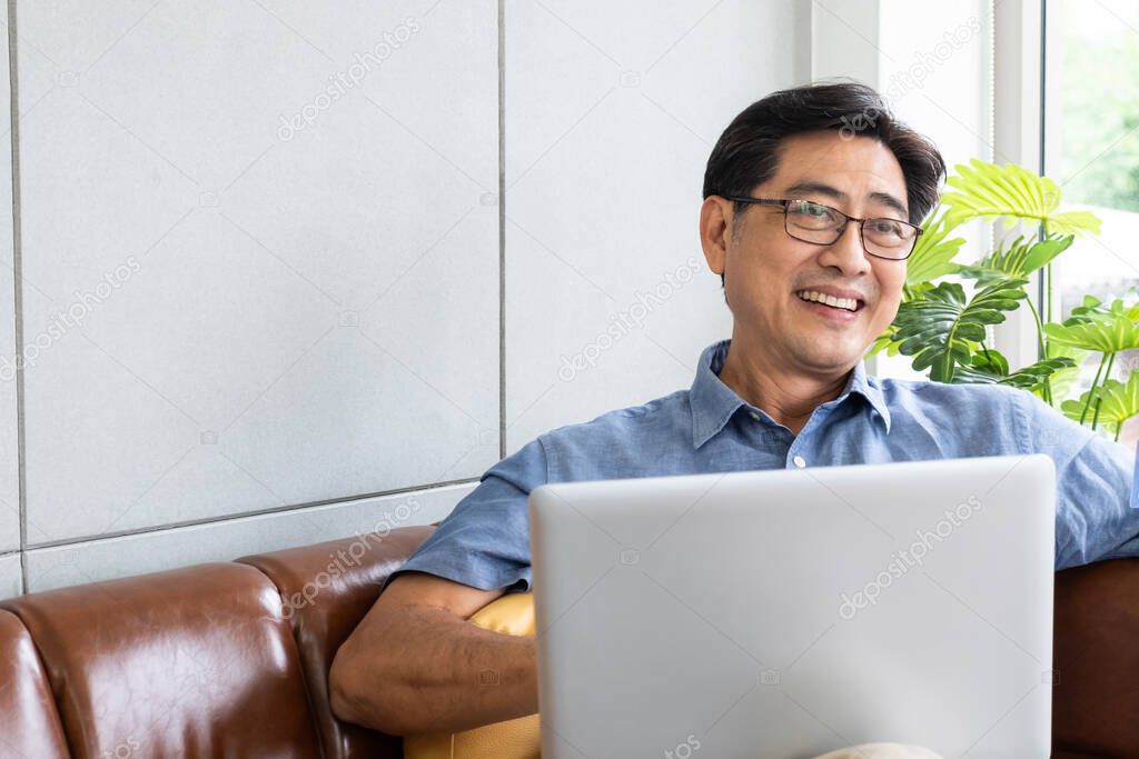 Indoor shot inside of the house of senior Asian men sit down on sofa and using computer laptop, browsing internet with smile on his face. Happy senior with technology and copy space for text.