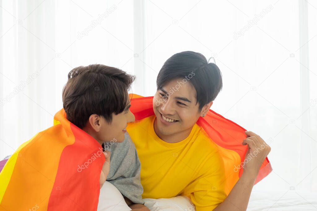 Selective focus at face. Handsome young Asian gay homosexual couple. Hug and embrace on the bed. With rainbow flag as LGBT sign. Gender equality and right. Romantic moment, same sex relationship.