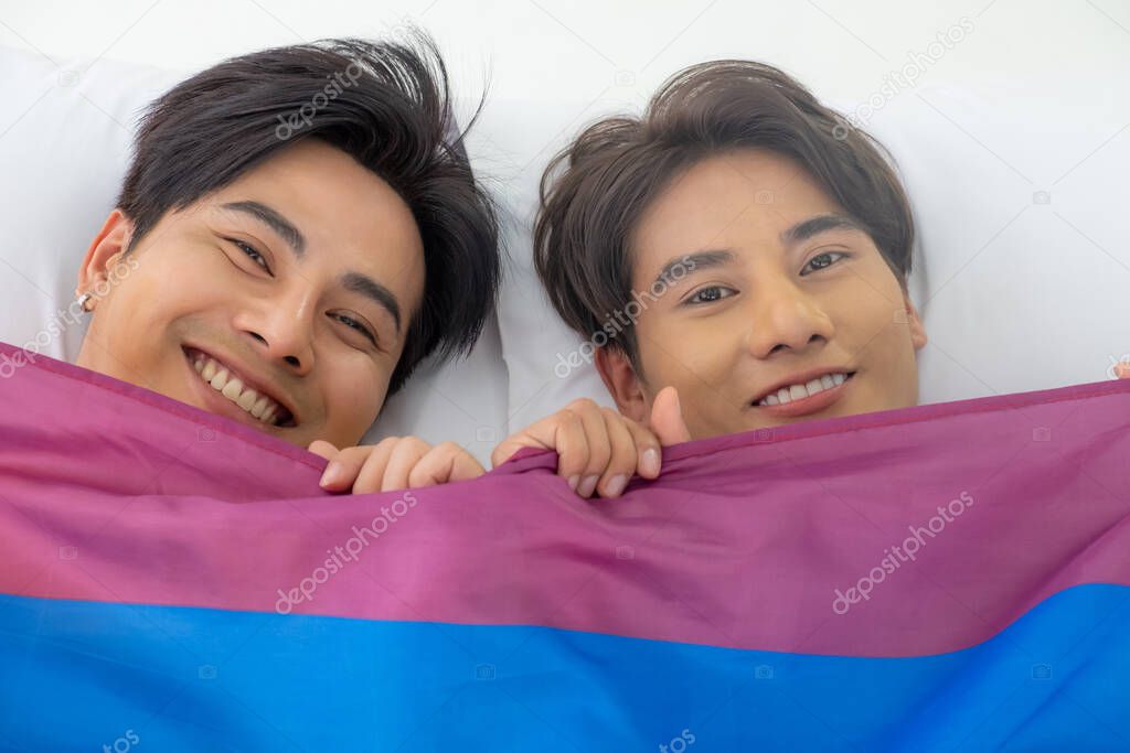 Selective focus at face. Handsome young Asian gay homosexual couple. Hug and embrace on the bed. With rainbow flag as LGBT sign. Gender equality and right. Romantic moment, same sex relationship.