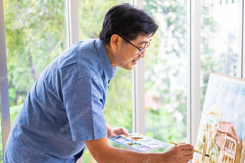 Indoor shot of senior Asian men using brush and oil color to paint on painting canvas. With smile on his face. Happy retirement concept for active senior. With Blurred green nature background.