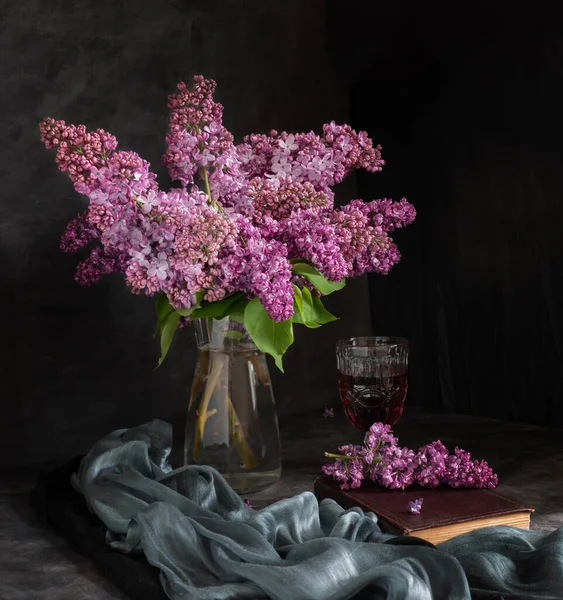 A bouquet of lilacs in a glass vase, a glass of red wine and a book on a dark background. Vintage still life