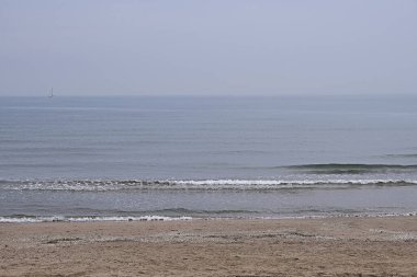 Sakin suları ve arka planında teknesi olan ıssız bir sahil, pastel tonlar, kum, bulutlu bir gün, kontrastsız