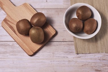 Tahta tahtada ve kasede birkaç kivi var. Tahta tahtalar, zenital manzara, boş alan, beyaz kase, kahverengi masa örtüsü..