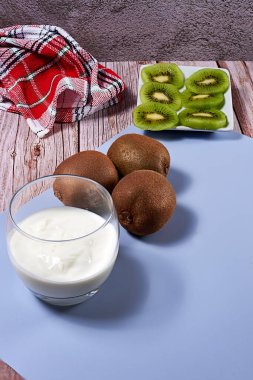 Bardakta yoğurt ve kırık kivi var. Ahşap zemin, ahşap tahta, bütün kivi, beyaz ve kırmızı mutfak havlusu, mavi paspas..