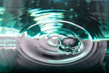 367.makro fotoğraf, bir su damlası tarafından oluşturulan dinamik sıçrama ve yayılan dalgaları yakalar. Görüntü, parlak, yansıtıcı renklerle, mavi-yeşil ve siyah renkler tarafından yönetilir.