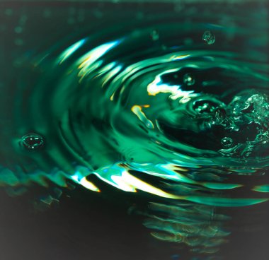 340 'lık damlacık suya çarpıp sıçrama ve eş merkezli dalgalanmalar oluşturur. Yeşil-sarı ışıklandırma ve yansımalar akışkan hareketi, yüzey gerilimi ve soyut bileşimi vurgular.