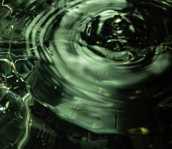 Su yüzeyinin 465 'lik yakın çekimi eşmerkezli dalgalanma halkaları ve yeşil yansımaları gösteriyor. Işıklandırma akışkan hareket ve dokuyu vurguluyor. Görüntü dalga davranışlarını, yüzey gerilimini ve optik bozulmayı yakalar.