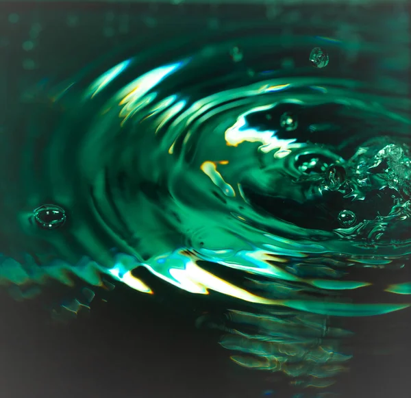 340 'lık damlacık suya çarpıp sıçrama ve eş merkezli dalgalanmalar oluşturur. Yeşil-sarı ışıklandırma ve yansımalar akışkan hareketi, yüzey gerilimi ve soyut bileşimi vurgular.