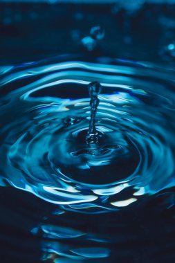 328.Damlacık mavi renkli suya çarpıp dikey sıçrama ve eş merkezli dalgalanmalar oluşturur. Işıklandırma akışkan hareketi, yüzey gerilimi ve dinamik görsel bileşimi vurgular.