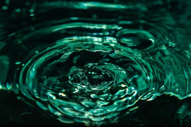 386.Su yüzeyine yakın çekim darbeden dolayı oluşan dalgalanma halkalarını gösteriyor. Mavi-yeşil ışık soyut dalga desenleri yaratır. Merkezi sıçrama sıvı hareketi ve yüzey gerilimi davranışını vurgular.