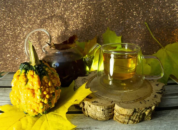 Chá de outono em uma caneca de vidro t um bule de vidro entre folhas amarelas de bordo e abóbora em um fundo borrado espumante, foco seletivo — Fotografia de Stock