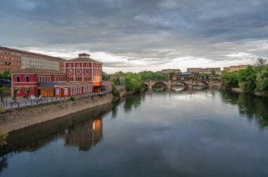 İspanya, La Rioja, Logrono 'daki Ebro Nehri' nin alacakaranlık manzarası nehir kenarındaki binalar ve sıcak akşam ışığı suyu yansıtıyor.