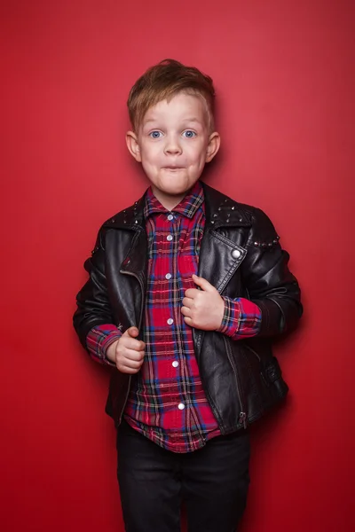 Mode liten pojke klädd i en skinnjacka. Studio porträtt över röd bakgrund — Stockfoto