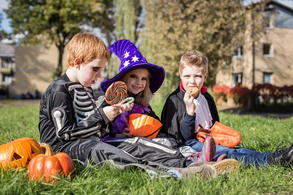 Tre piccoli amici carini seduti sull'erba e mangiare caramelle di Halloween — Foto Stock