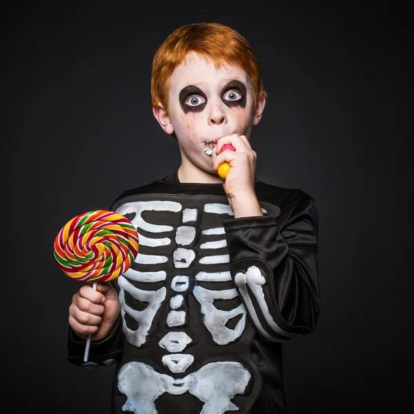 Joyeux jeune garçon aux cheveux roux avec costume squelette tenant et mangeant des bonbons colorés — Photo
