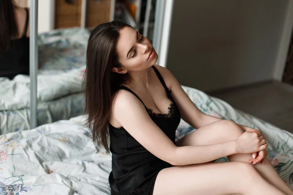 Vacker ung kvinna i en svart nattlinne sitter på sängen. — Stockfoto