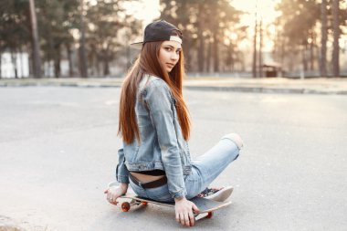 Şık giyim günbatımında bir kaykay üzerinde oturan genç güzel moda kız
